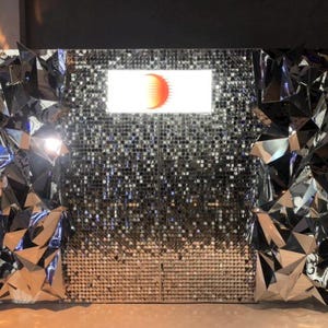 May include: A silver mirrored backdrop with a geometric design. A screen with a red sun graphic is in the center of the backdrop.