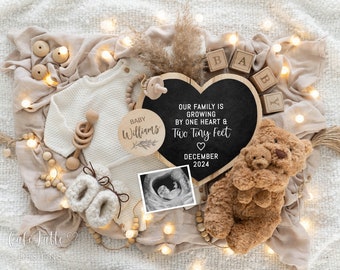 May include: A heart-shaped chalkboard sign with the text "Our family is growing by one heart & two tiny feet" and the date "December 2024". The sign is surrounded by a white knitted sweater, a wooden baby rattle, a wooden circle with the text "Baby Williams", a teddy bear, and a string of lights.