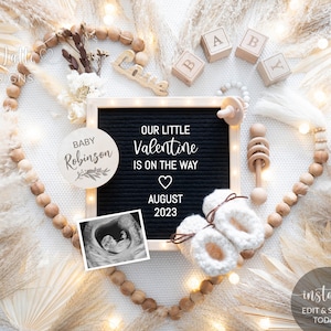 May include: A black letter board with the text "Our Little Valentine is on the way" and "August 2023". There is a wooden circle with the text "Baby Robinson" and a small ultrasound image. The letter board is surrounded by wooden beads, a wooden rattle, and a pair of white crocheted baby booties.