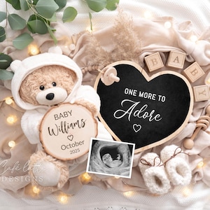 May include: A white teddy bear wearing a white hooded outfit holds a wooden sign that reads "Baby Williams October 2023". A black chalkboard heart with the text "One More to Adore" is in the background. The image is surrounded by a white blanket with wooden blocks that spell out "Baby".