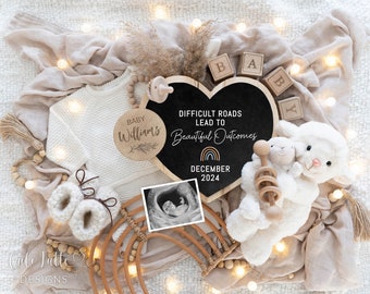 May include: A heart-shaped chalkboard sign with the text "Difficult roads lead to beautiful outcomes" and "December 2024".  The sign is surrounded by a white knitted blanket, wooden blocks spelling "BABY", a wooden circle with the text "Baby Williams", a baby rattle, a pair of knitted booties, and a framed ultrasound image.