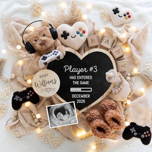 May include: A baby announcement featuring a teddy bear with headphones and a game controller. A heart-shaped chalkboard reads "Player #3 has entered the game" with the due date. Other items include baby booties, a dummy, and wooden blocks spelling "BABY".