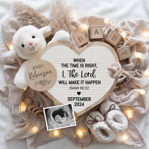 May include: A heart-shaped wooden sign with the text "WHEN THE TIME IS RIGHT, 1. THE LORD WILL MAKE IT HAPPEN ISAIAH 60:22 SEPTEMBER 2024". The sign is surrounded by a white stuffed lamb, a wooden baby block that says "BABY", a wooden circle with the text "BABY Robinson", a pacifier, a pair of white crocheted baby booties, and a string of fairy lights.