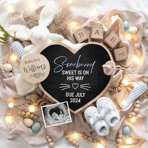 May include: A heart-shaped chalkboard sign with the text "Somebunny Sweet is on His Way" and "Due July 2024". The sign is surrounded by a white bunny, wooden blocks spelling "BABY", a blue and white gingham ribbon, and a string of fairy lights.