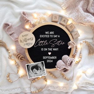 May include: A black letter board with the text "We are excited to say a little sister is on the way!" and the date "September 2024". There is a small ultrasound image, a wooden baby rattle, a pair of pink baby booties, and a wooden circle with the text "Baby Williams" on it.