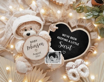 Könnte beinhalten: Ein herzförmiges Schild aus Holz mit dem Text "We've been keeping a little secret" in weißen Buchstaben. Ein Teddybär, der ein weißes Kapuzenoutfit trägt, hält ein rundes Schild aus Holz mit dem Text "Baby Robinson" und "August 2023". Ein Schwarzweiß-Ultraschallbild befindet sich im Hintergrund.
