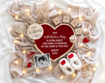 Op de afbeelding: Een rood hartvormig houten bord met de tekst "This Valentine's Day is extra sweet our family is growing by one heart & two feet August 2024". Het bord is omringd door een wit deken, een houten babyblok met de tekst "Baby", een houten cirkel met de tekst "Baby Williams", een kleine echografiefoto, een paar witte gehaakte slofjes met rode hartjes en een houten kralenketting.