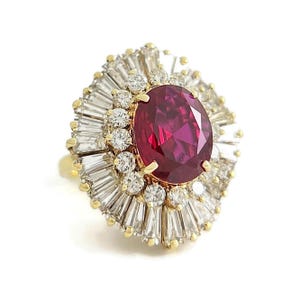 Puede incluir: Anillo de oro con un gran rubí ovalado en el centro. El rubí está rodeado por un halo de diamantes redondos más pequeños y diamantes baguette, creando un efecto de sol. El anillo tiene un diseño clásico y elegante.