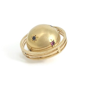May include: A gold-colored ring with a spherical center, adorned with small, star-shaped gems in blue and red. The ring features multiple bands encircling the sphere, creating a celestial or planetary design. The ring is set against a white background.