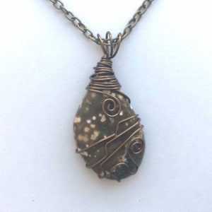 May include: A handcrafted pendant necklace featuring a teardrop-shaped stone wrapped in copper wire. The stone has a speckled pattern of green and white. The necklace chain is a matching antique bronze color.