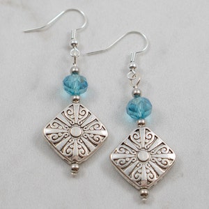 May include: Silver earrings with a square pendant featuring a detailed design and a light blue crystal bead.