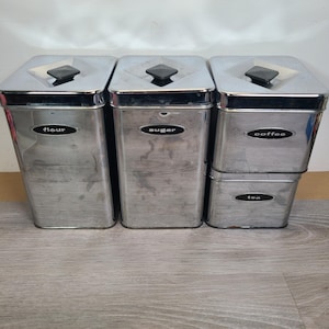 Könnte beinhalten: Ein Satz von vier Küchenbehältern aus Edelstahl. Die Behälter sind mit "Mehl", "Zucker", "Kaffee" und "Tee" beschriftet. Jeder Behälter hat einen rautenförmigen Deckel mit einem schwarzen Knopf.