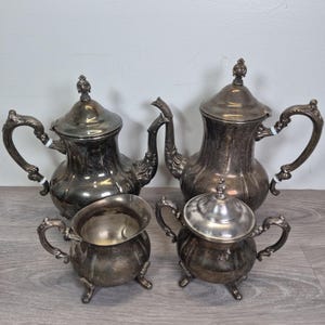 May include: A four-piece silver-plated tea set consisting of two teapots with lids, a creamer, and a lidded sugar bowl. Each piece features a bulbous body with vertical fluting, ornate curved handles, and decorative finials on the lids.