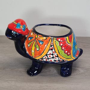 May include: A ceramic turtle planter with a vibrant, hand-painted design. The turtle is primarily navy blue with a red hat and colourful accents of orange, yellow, green, and blue. The planter has a large opening for plants.