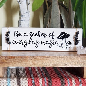 May include: White wooden sign with the text "Be a seeker of everyday magic" and a black and white mushroom illustration. The sign is decorated with black stones and twine.