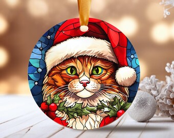 May include: A round Christmas ornament featuring a cat wearing a Santa hat and holding a sprig of holly with red berries. The cat has green eyes and orange and white fur. The ornament is made of glass and has a stained glass look.