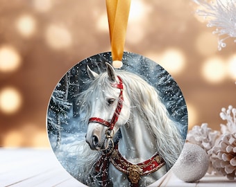 May include: Round Christmas ornament featuring a white horse with a red bridle and harness, set against a snowy forest backdrop. The ornament has a gold ribbon for hanging. A silver ball and pinecone are in the background.