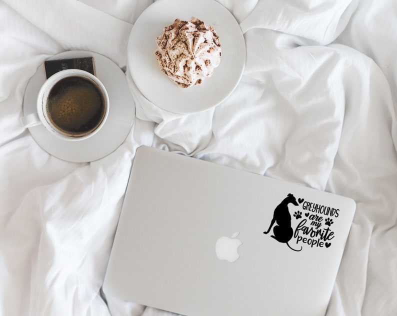 Peut inclure: Un ordinateur portable blanc avec une silhouette noire de l&eacute;vrier et le texte "Les l&eacute;vriers sont mes personnes pr&eacute;f&eacute;r&eacute;es" avec des empreintes de pattes et des c&oelig;urs. L'ordinateur portable est sur un couvre-lit blanc avec une tasse de caf&eacute; et une p&acirc;tisserie.