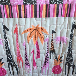 May include: A pink, orange and black quilted baby blanket featuring a giraffe and palm tree design. The blanket has a geometric pattern on the bottom border.