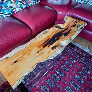 May include: A natural wood coffee table with a live edge design. The table has a dark brown stain in the center and is supported by a black metal base. The table is sitting on a red and black patterned rug.