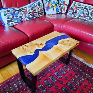 May include: A rectangular coffee table with a natural wood top and a blue epoxy river design. The table has black metal legs and sits on a red patterned rug. The table's dimensions are approximately 48 inches long and 24 inches wide.