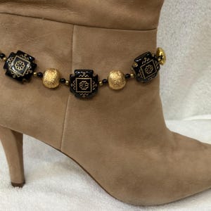 May include: A boot accessory chain featuring alternating black square beads with gold floral patterns and textured gold spherical beads. The chain is secured with a gold-toned metal clasp and small black spacer beads, wrapped around a tan suede boot.