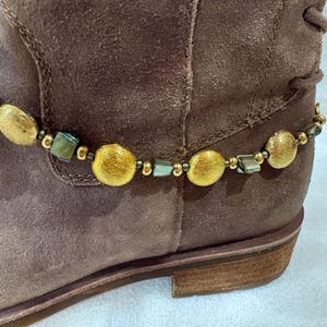 May include: A brown suede boot with a decorative ankle bracelet. The bracelet features gold-colored, round beads and smaller, square, iridescent beads strung together with a gold-colored chain. The boot has a low heel.