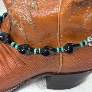 Op de afbeelding: Een bruine leren cowboylaars met een decoratieve enkelband. De armband heeft zwarte, turquoise en zilveren kralen. De laars heeft decoratieve stiksels en een textuur.