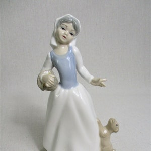 May include: A porcelain figurine of a girl in a white dress and blue bodice, holding a basket and walking with a small brown dog.