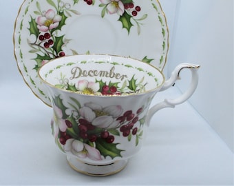 May include: A white teacup and saucer set with a floral design featuring pink flowers, red berries, and green leaves. The word "December" is written in gold script on the cup. The set has gold trim.