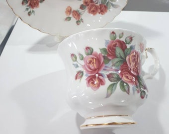 Puede incluir: Juego de taza y platillo blancos con diseño floral de rosas. La taza tiene un asa y un ribete dorado. El platillo tiene un borde festoneado con ribete dorado. Las rosas son rojas y rosas con hojas verdes.