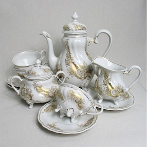 Puede incluir: Un juego de té de porcelana blanca con detalles dorados. El juego incluye una tetera, una jarra de crema, un azucarero y dos tazas de té y platillos. Las tazas de té y los platillos tienen un borde festoneado y la tetera tiene una tapa con un pomo.