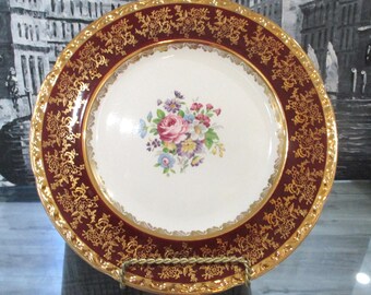 May include: A vintage decorative plate with a white center featuring a floral bouquet of pink, blue, and yellow flowers. The plate has a burgundy border with gold filigree and a gold rim. The plate is displayed on a gold-colored stand.