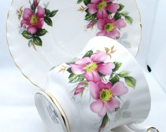 Puede incluir: Un juego de taza y plato de porcelana blanca con motivos florales de rosas silvestres rosas, hojas verdes y centros amarillos. Los bordes de la taza y el plato tienen un acabado dorado. La base de la taza muestra la marca Royal Albert Prairie Rose.