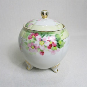 May include: A white porcelain cookie jar with a lid and three feet. The jar is decorated with pink and red flowers and green leaves. The lid has a gold knob.