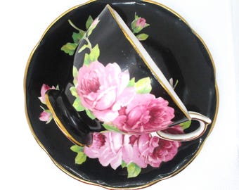 May include: Black teacup and saucer set with pink rose floral design. The cup and saucer have a gold trim. The teacup is tilted, revealing the white interior. The set is likely vintage or antique.