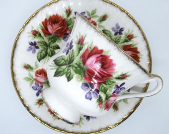 May include: White teacup and saucer set with a floral rose design in red, pink, and purple. The teacup and saucer have a gold trim. The teacup is resting on the saucer.