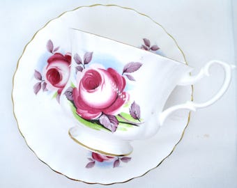 May include: A white teacup and saucer with a pink rose floral pattern. The cup has a delicate handle and a gold rim.