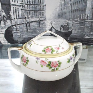 May include: White porcelain sugar bowl with lid, featuring a floral design with pink and purple flowers and green leaves. The bowl has a gold trim and a handle on the lid.