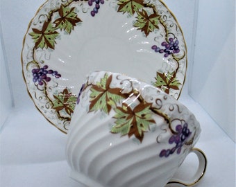 May include: A white teacup and saucer set with a grapevine design. The cup and saucer are decorated with purple grapes, green leaves, and gold accents.