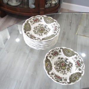 Può includere: Una pila di otto ciotole rotonde in ceramica e una ciotola singola con bordo smerlato. Le ciotole bianche sono decorate con un dettagliato motivo floreale nei toni del verde oliva, rosso, giallo e blu, con un motivo botanico centrale.