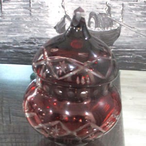 Pode incluir: Um frasco de vidro lapidado vermelho rubi com tampa. O frasco tem um corpo redondo e uma tampa em cúpula, ambos decorados com padrões de estrelas e diamantes. O frasco está sobre uma superfície refletora, com uma pintura de um canal veneziano ao fundo.