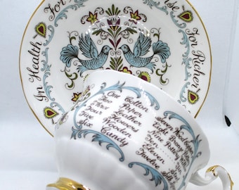 May include: A white porcelain teacup and saucer set with gold trim. The saucer features blue birds, floral motifs, and the text For Better, For Worse, In Health, For Richer. The cup lists anniversary themes from One Cotton to Twelve Silk Linen.