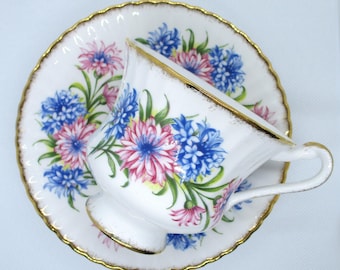 May include: A white teacup and saucer with a floral pattern. The pattern features pink and blue flowers with green leaves. The cup and saucer have a gold trim.