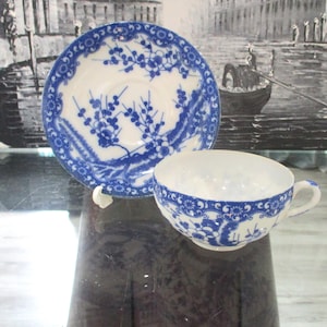 May include: A vintage teacup and saucer set. The white porcelain cup and saucer feature a blue floral design. The saucer has a scalloped edge. The cup has a handle and a matching blue floral pattern around the rim.