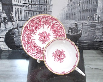 Può includere: Un set di tazza e piattino bianchi con un motivo floreale rosso e bordi dorati. La tazza ha un manico e il piattino è rotondo. Il set è esposto su una superficie scura.