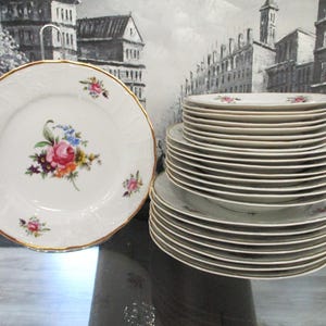 May include: A set of white porcelain plates with a floral design. The plates are stacked in a pile, with the top plate featuring a pink rose and other flowers. The plates have a gold rim.