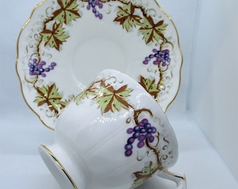 May include: A white teacup and saucer with a gold rim and a grapevine design. The grapes are purple and the leaves are green and brown.