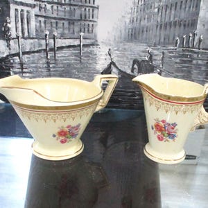 Op de afbeelding: Een vintage roomkleurige roomkan en suikerset met gouden rand en bloemmotieven. De roomkan heeft een brede, ondiepe kom en een handvat. De suikerpot is hoger en heeft een traditionelere vorm.