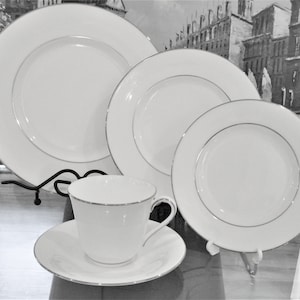 May include: A white porcelain dinner service with a silver trim. The set includes a dinner plate, salad plate, bread plate, and a teacup with saucer. The plates are displayed on black metal stands, and the teacup is on a glass surface.
